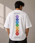 A man viewed from behind wearing a white oversized T-shirt with a vertical multicolor chakra graphic and text on the back, paired with dark pants, against a neutral indoor wall.