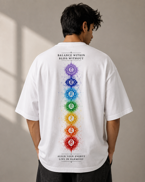 A man viewed from behind wearing a white oversized T-shirt with a vertical multicolor chakra graphic and text on the back, paired with dark pants, against a neutral indoor wall.